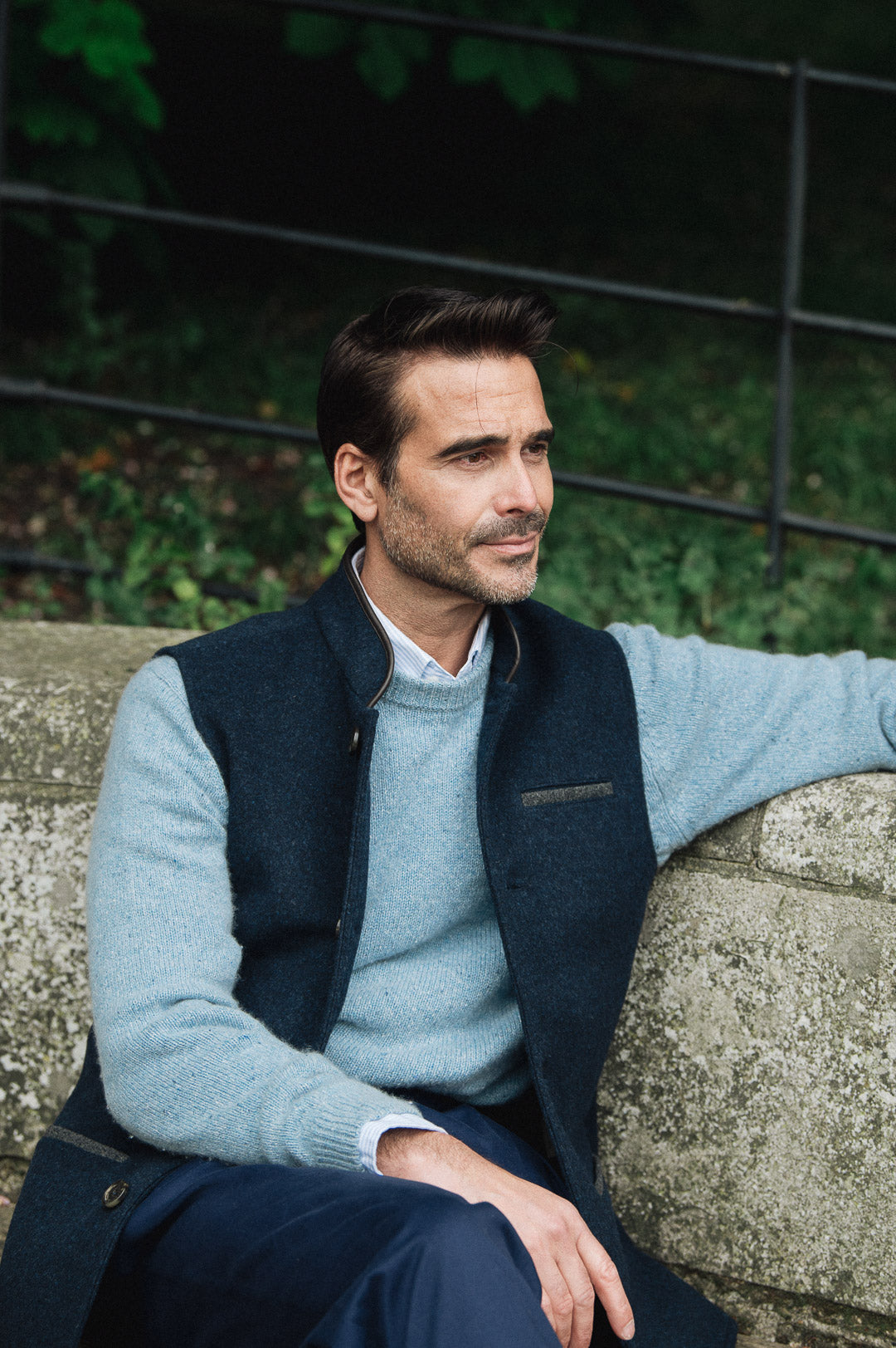Man wearing a Darzi Midnight Blue merino wool waistcoat sitting on a stone wall with a blurred background