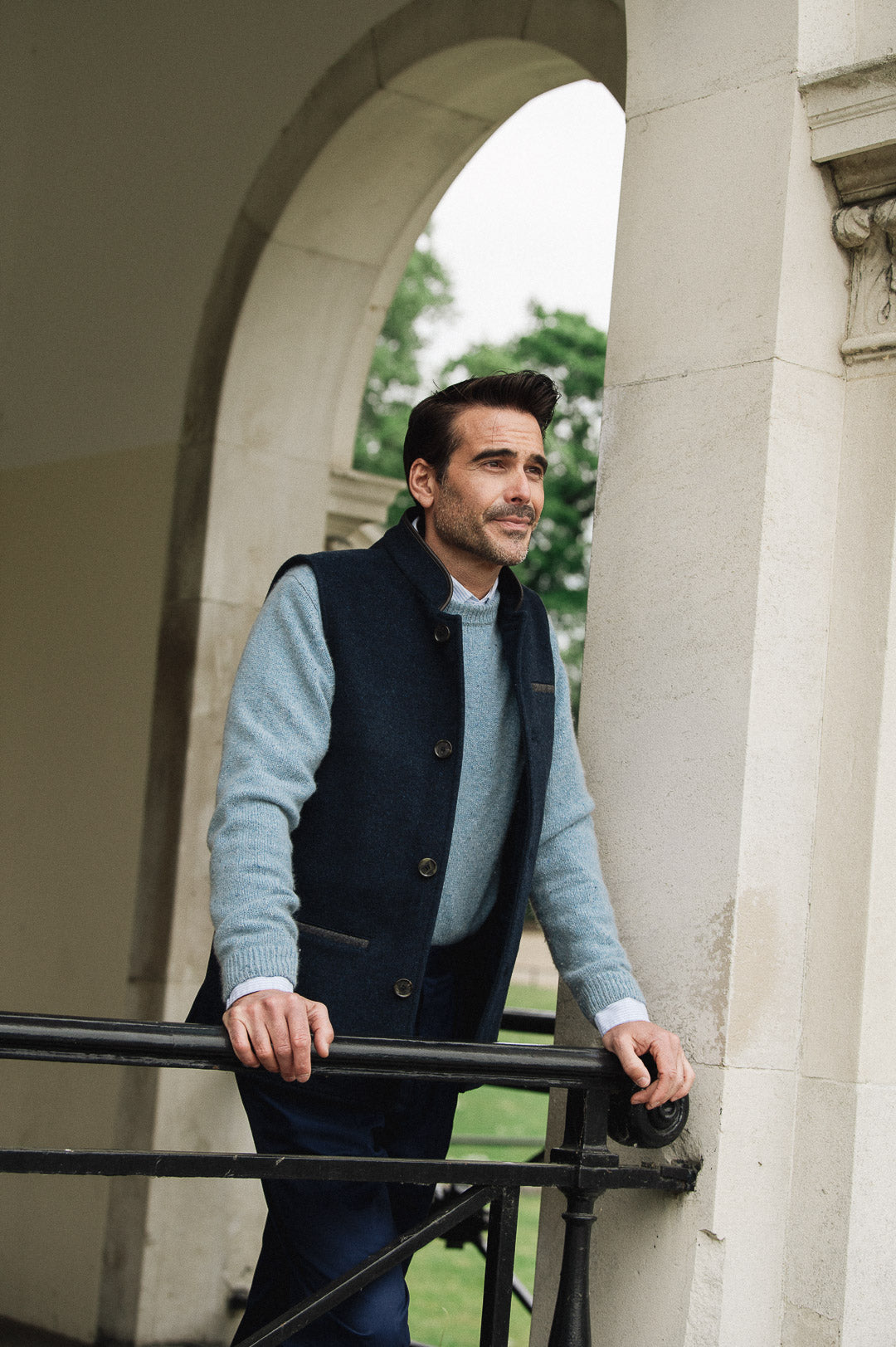 Man standing outdoors near a stone archway wearing a blue sweater and Darzi midnight blue merino vest
