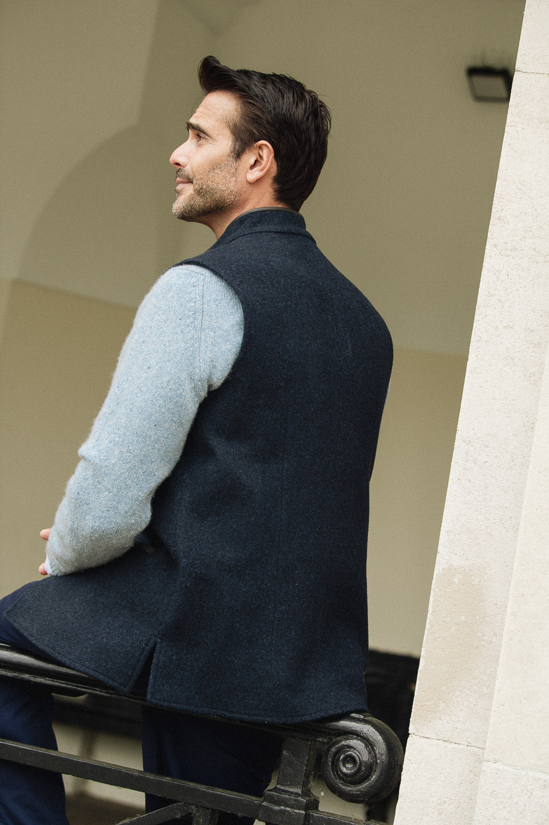 Man wearing a Darzi Midnight Blue merino wool waistcoat, sitting on a black railing - back detail