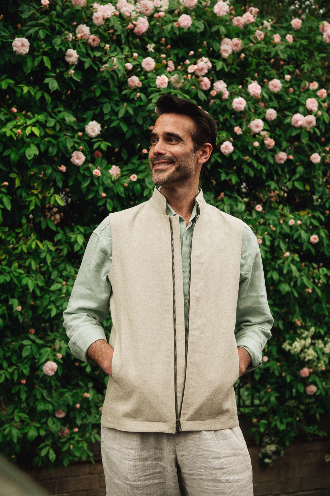 Man wearing a Darzi limestone linen Hillgate gilet standing in front of a rose bush