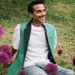Man sitting on grass with purple flowers, wearing a jade green Darzi linen waistcoat over a white shirt.