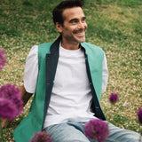 Man sitting on grass with purple flowers, wearing a jade green Darzi linen waistcoat over a white shirt.