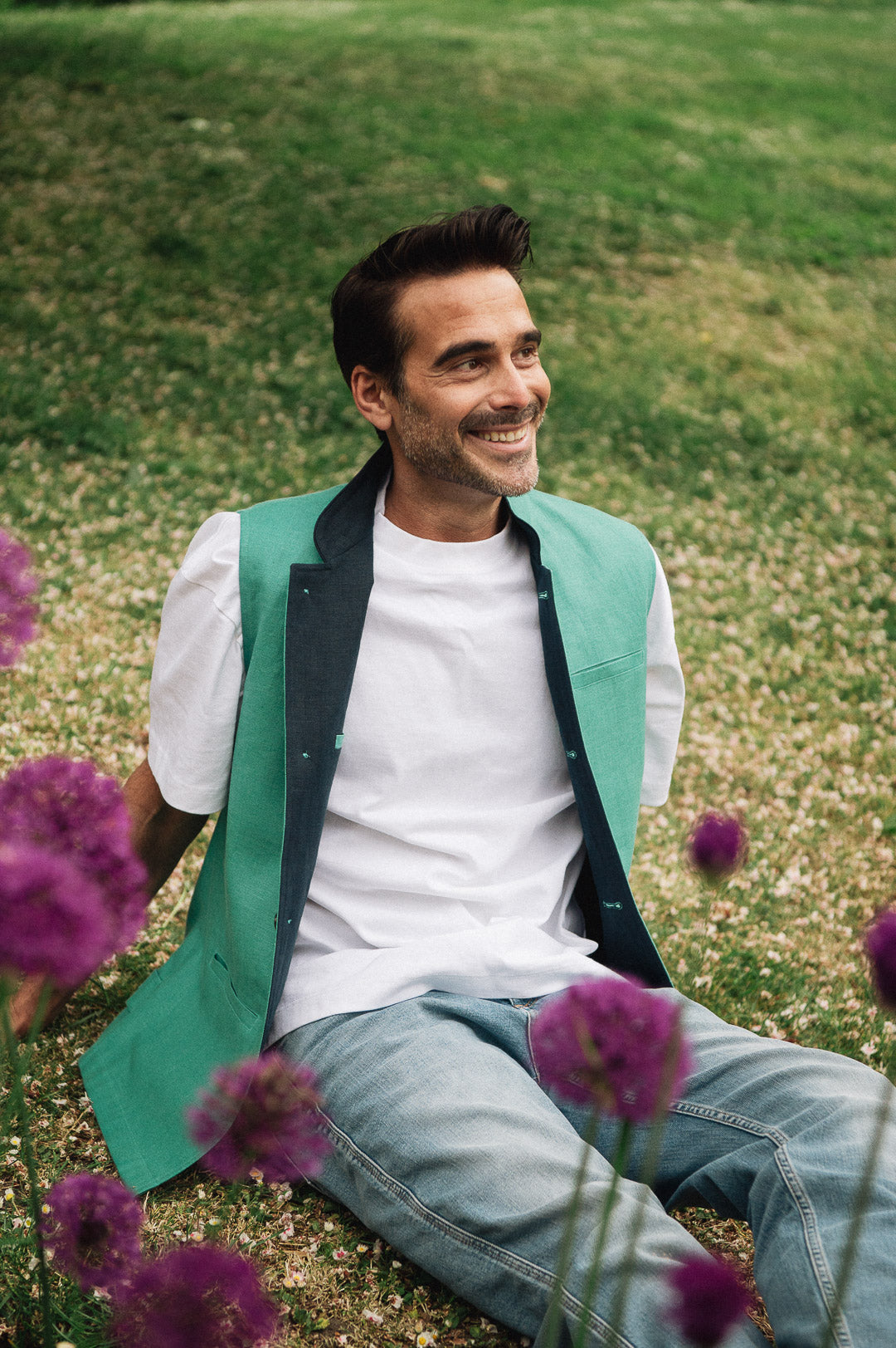 Man sitting on grass with purple flowers, wearing a jade green Darzi linen waistcoat over a white shirt.