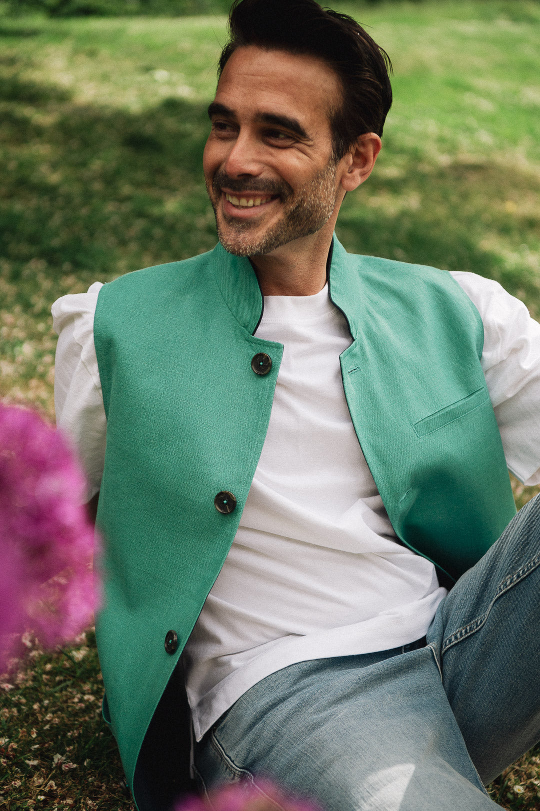 Man wearing a green Darzi linen vest over a white shirt, sitting outdoors with grass and flowers in the background.
