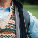 Person wearing a patterned jumper over a light blue shirt, under a Darzi navy linen Hillgate gilet with a blurred green background