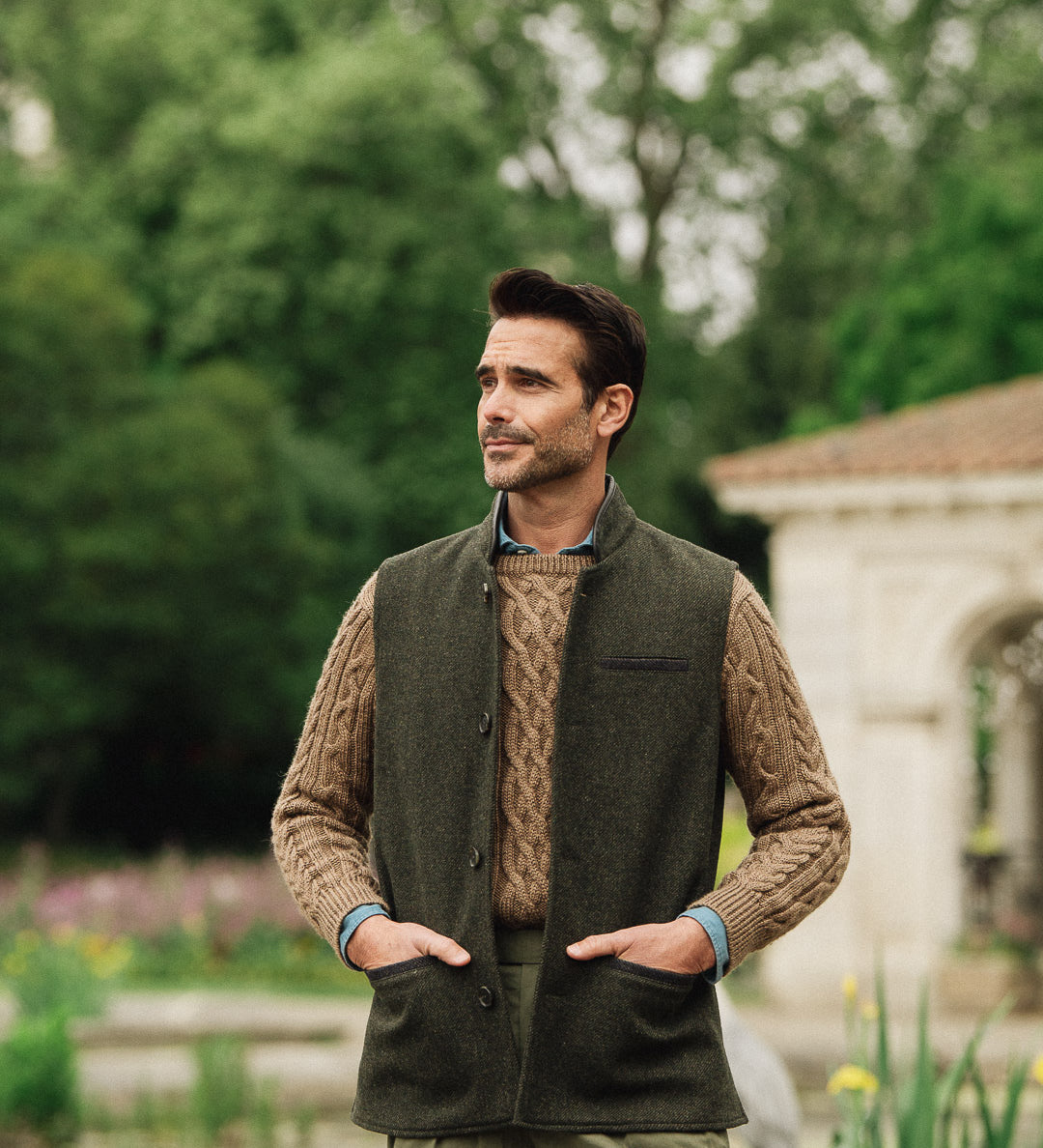 Man wearing a green wool vest over a brown sweater in an outdoor setting with trees and a building