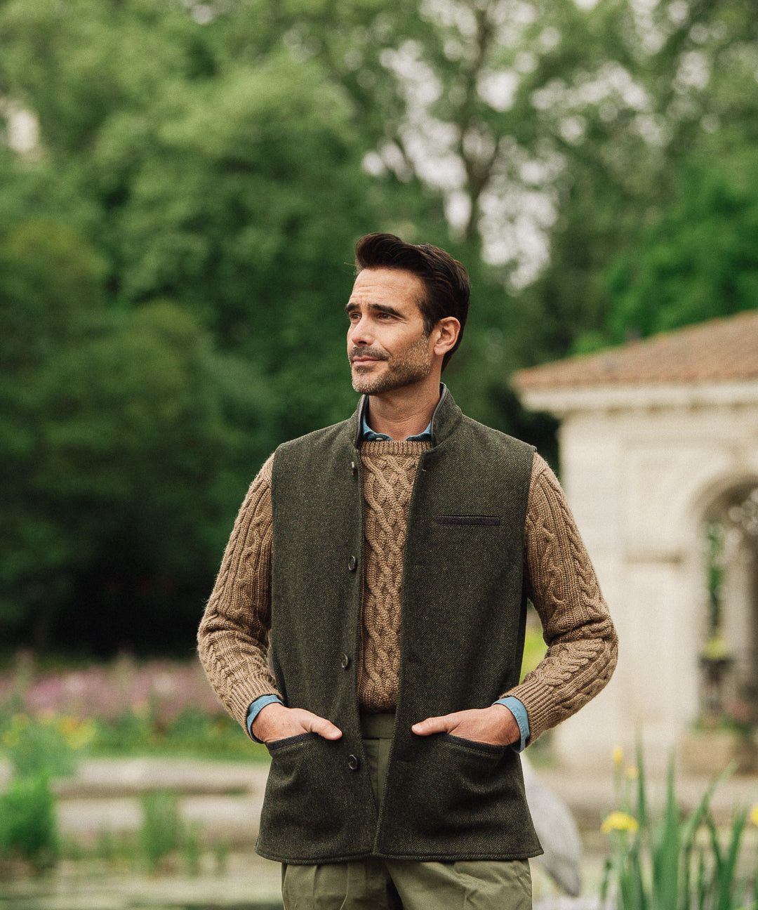 Man wearing a green wool vest over a brown sweater in an outdoor setting with trees and a building