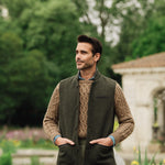 Man wearing a green wool vest over a brown sweater in an outdoor setting with trees and a building