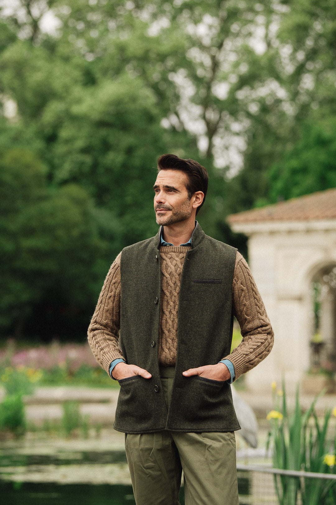 Man wearing a green wool vest over a brown sweater in an outdoor setting with trees and a building