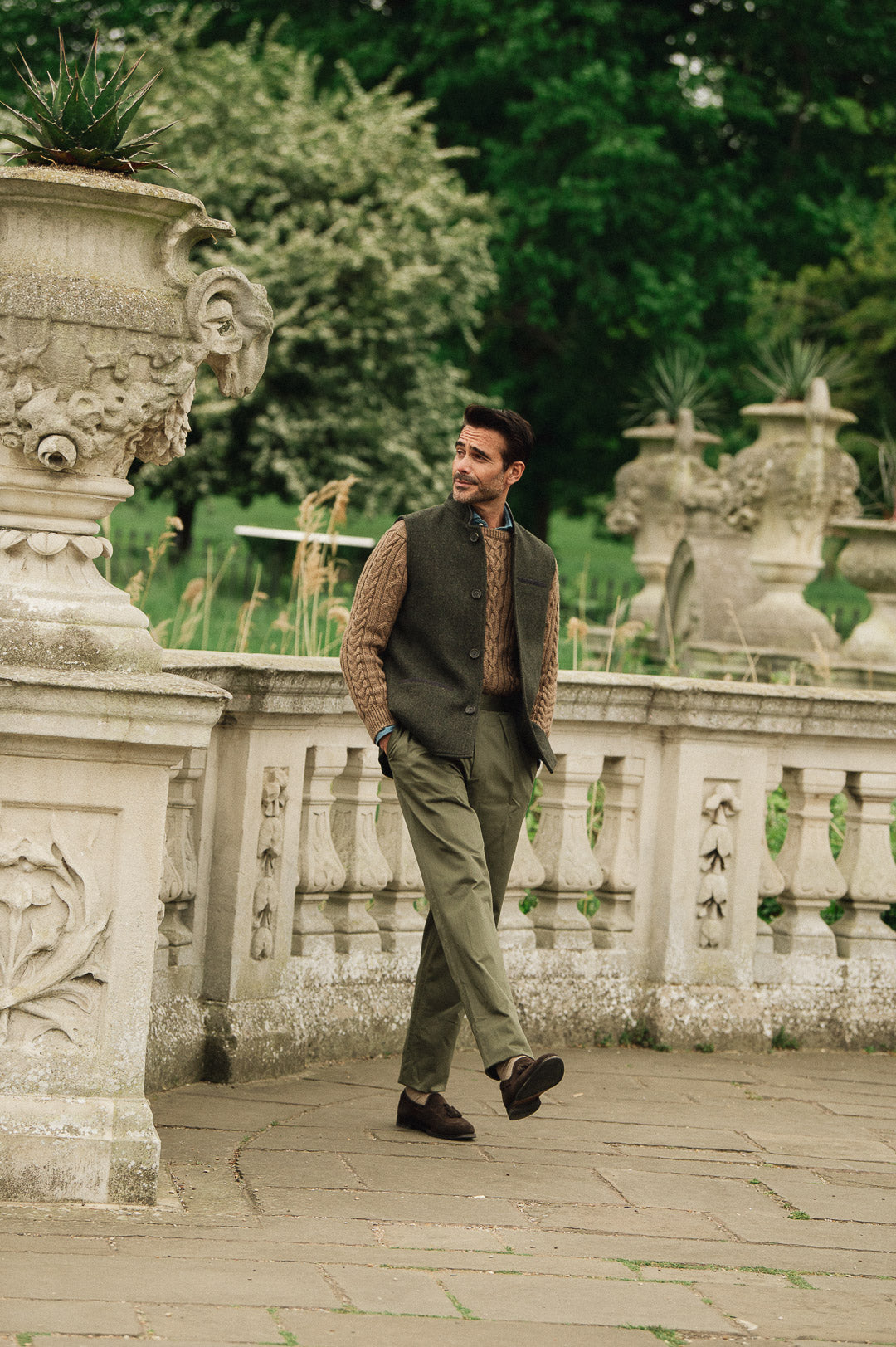 Man walking in a garden wearing a Darzi Helmsley wool waistcoat surrounded by Italianate  sculptures and greenery