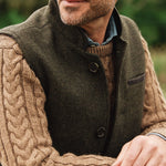 Man wearing a green brown Darzi wool vest over a dark sweater with a blurred natural background