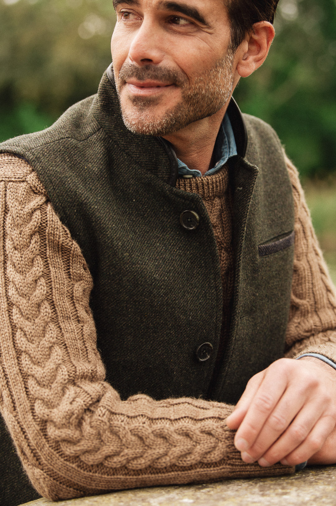 Man wearing a green brown Darzi wool vest over a dark sweater with a blurred natural background