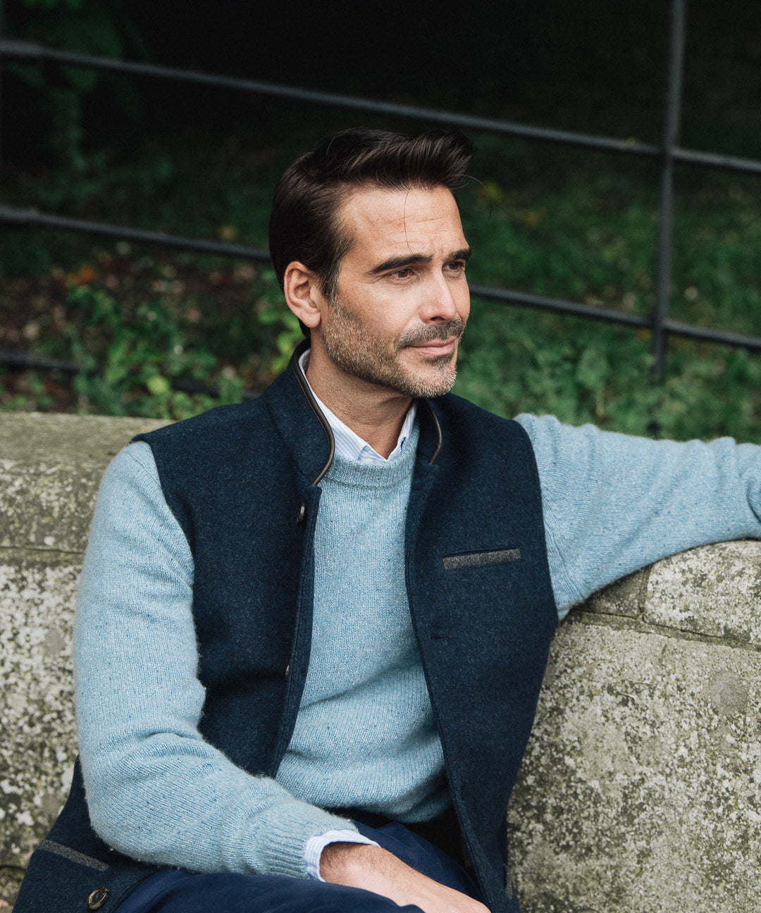 Man wearing a Darzi Midnight Blue merino wool waistcoat sitting on a stone wall with a blurred background