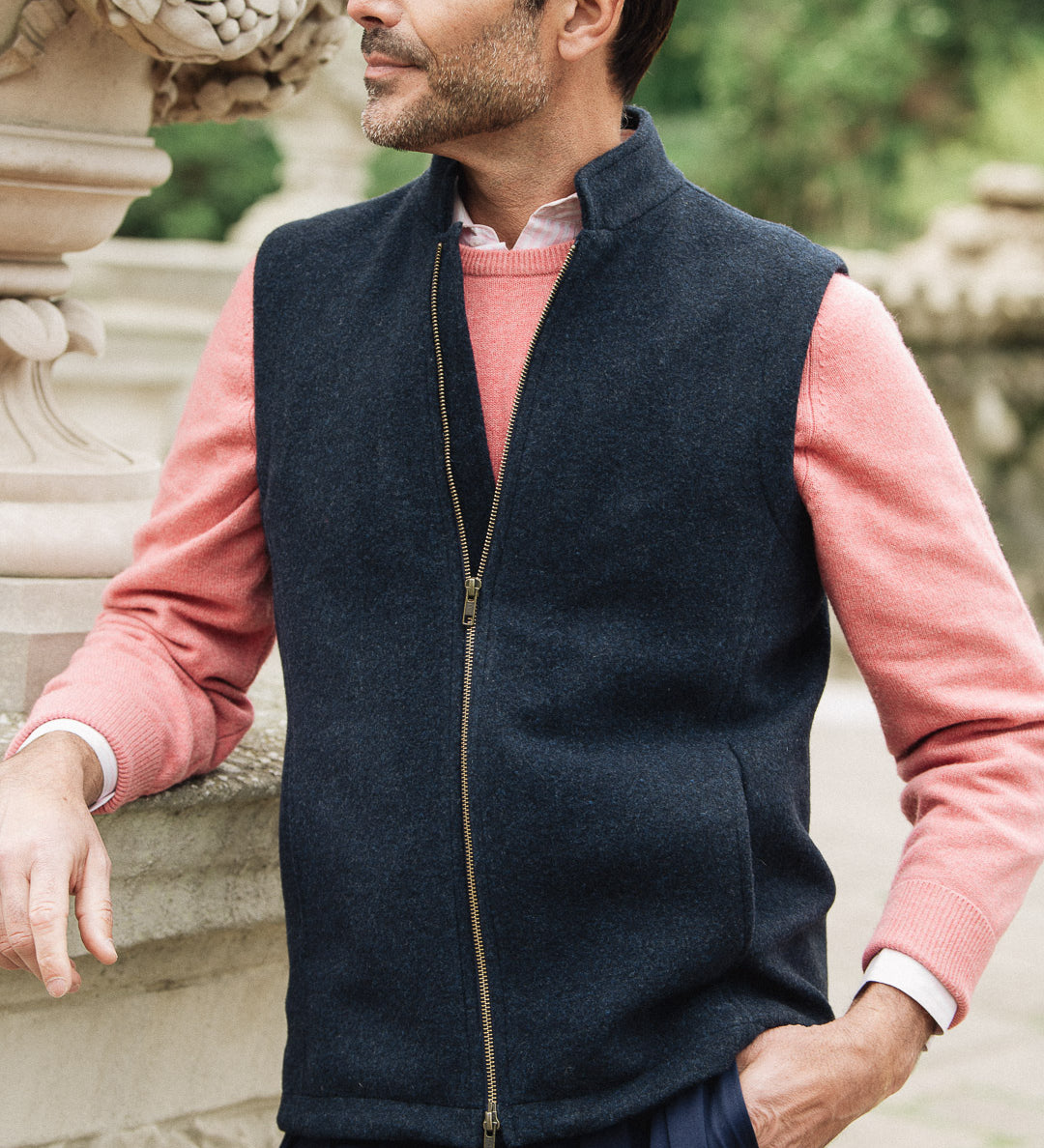Man wearing a navy Darzi merino wool gilet over a pink sweater with a blurred natural background