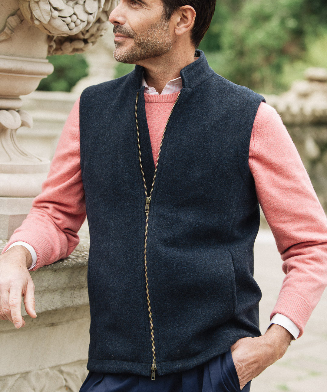 Man wearing a navy Darzi merino wool gilet over a pink sweater with a blurred natural background