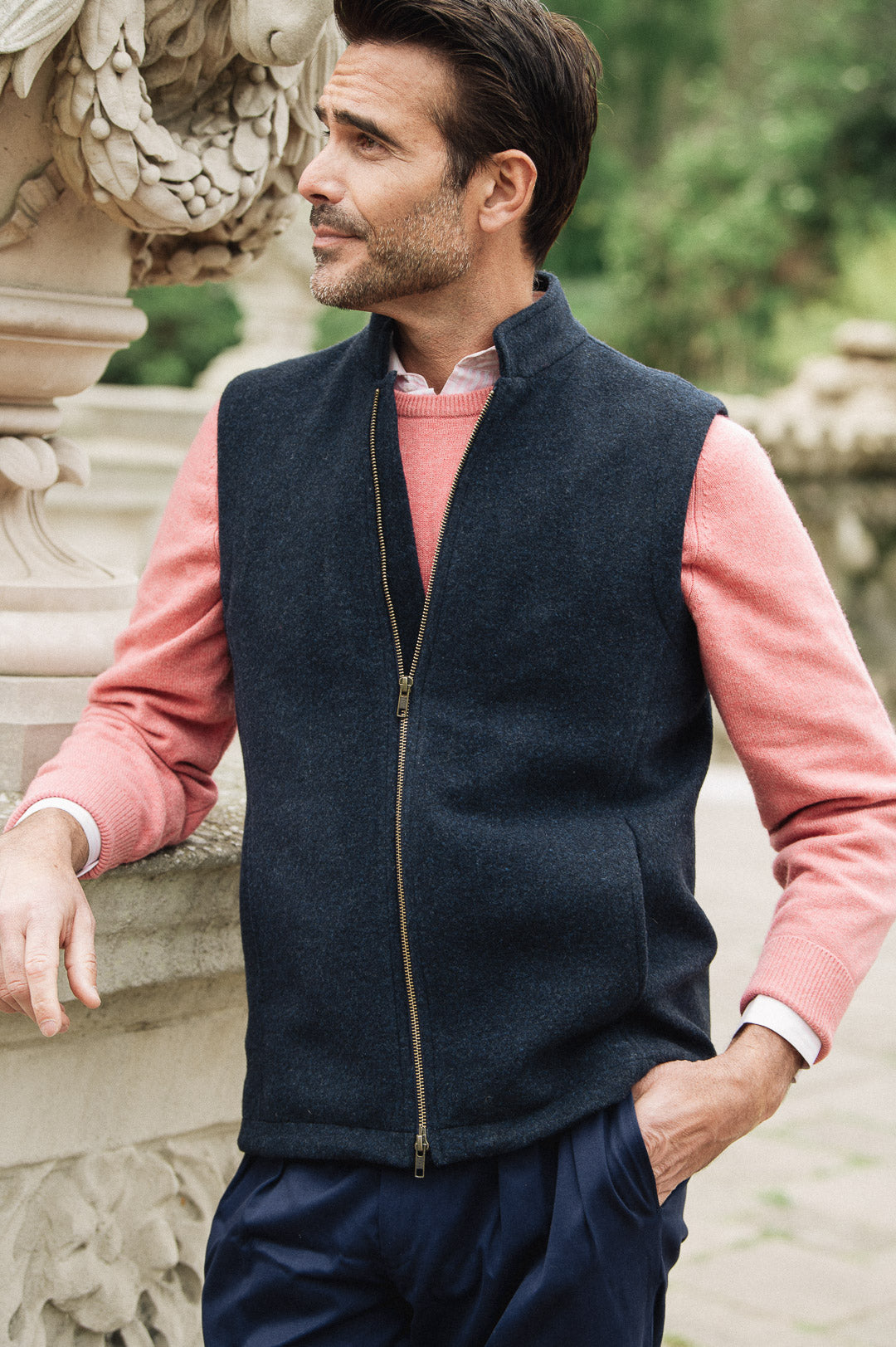 Man wearing a navy Darzi merino wool gilet over a pink sweater with a blurred natural background