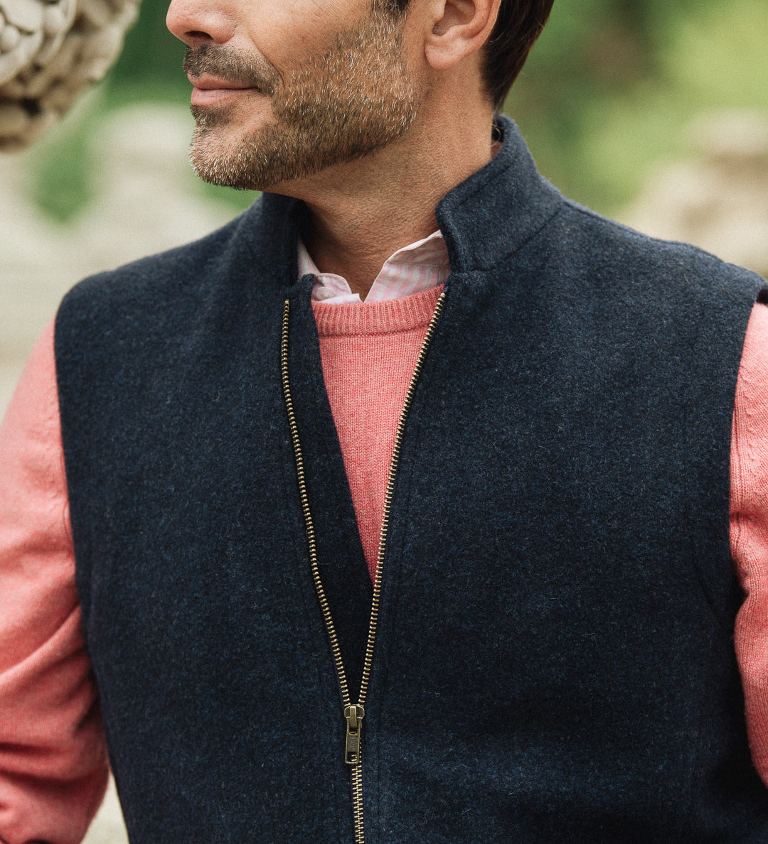 Man wearing a navy Darzi merino wool gilet over a pink sweater with a blurred natural background