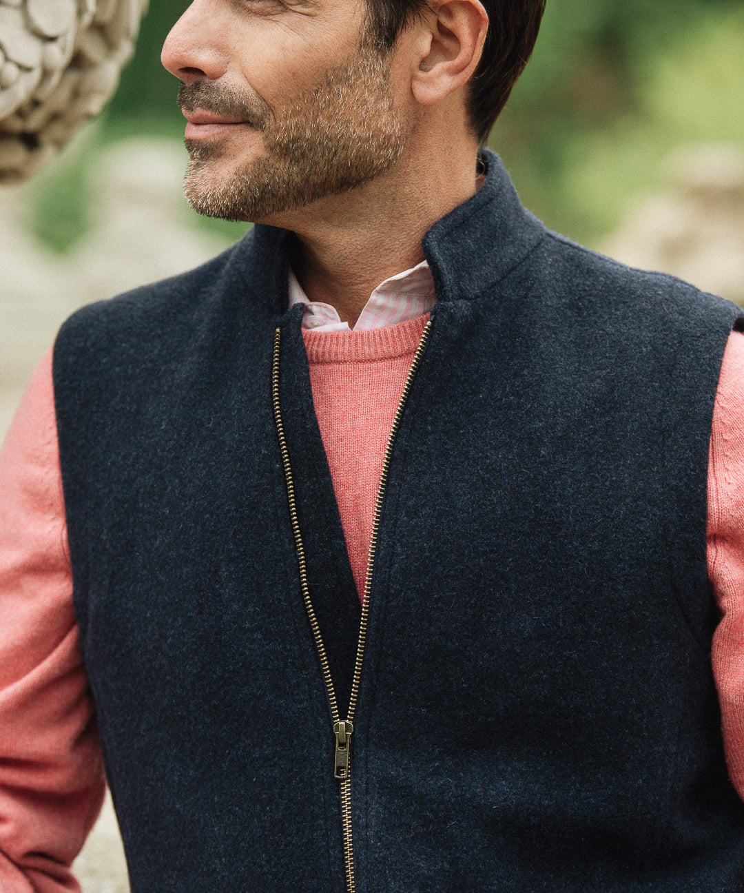 Man wearing a navy Darzi merino wool gilet over a pink sweater with a blurred natural background