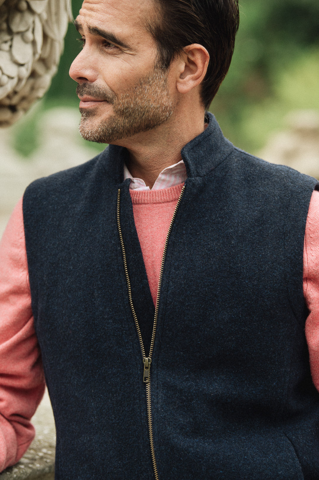 Man wearing a navy Darzi merino wool gilet over a pink sweater with a blurred natural background