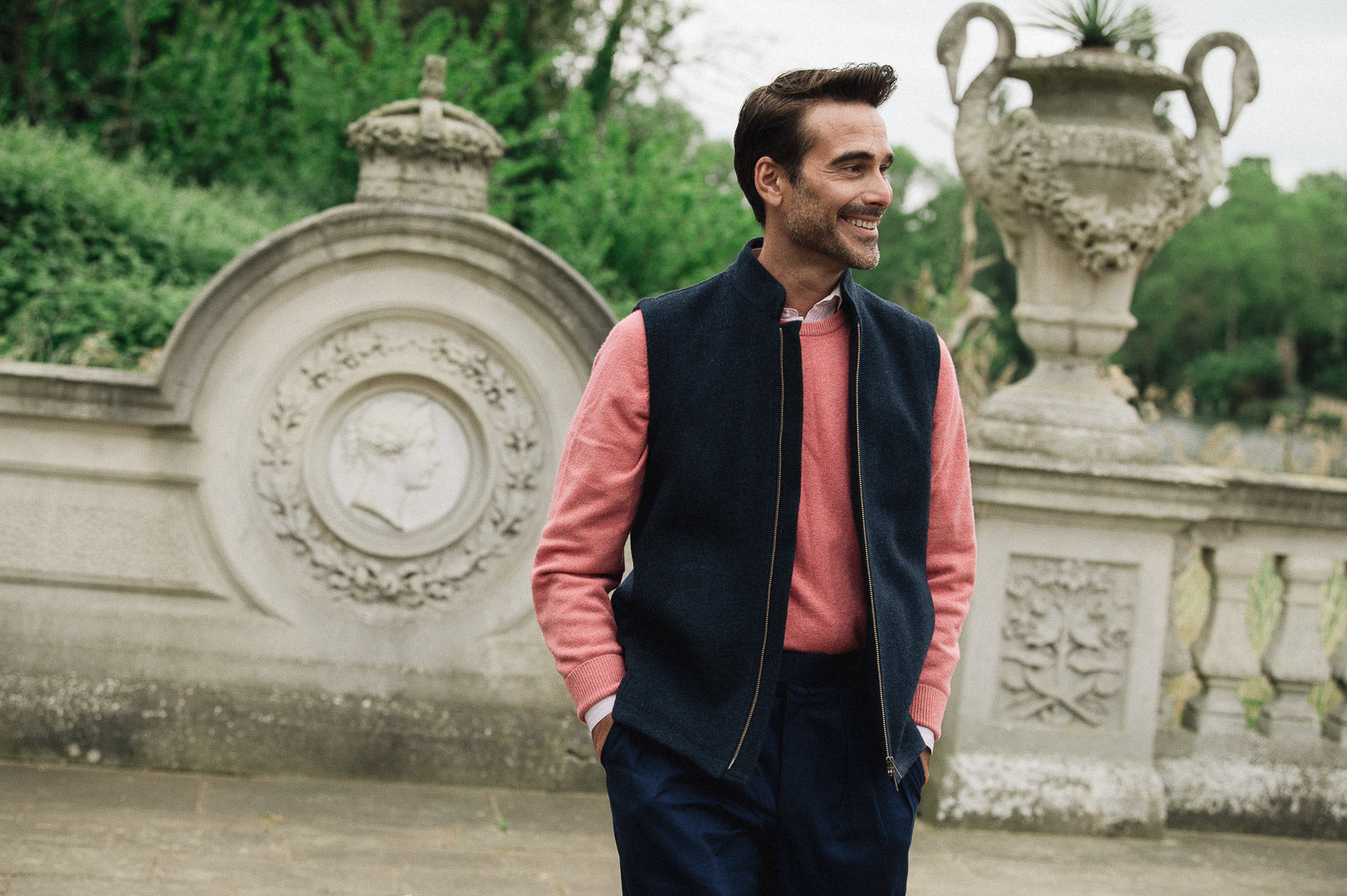 Man wearing a Darzi Hillgate heavyweight midnight blue merino wool gilet over a pink sweater in an Italian style garden setting