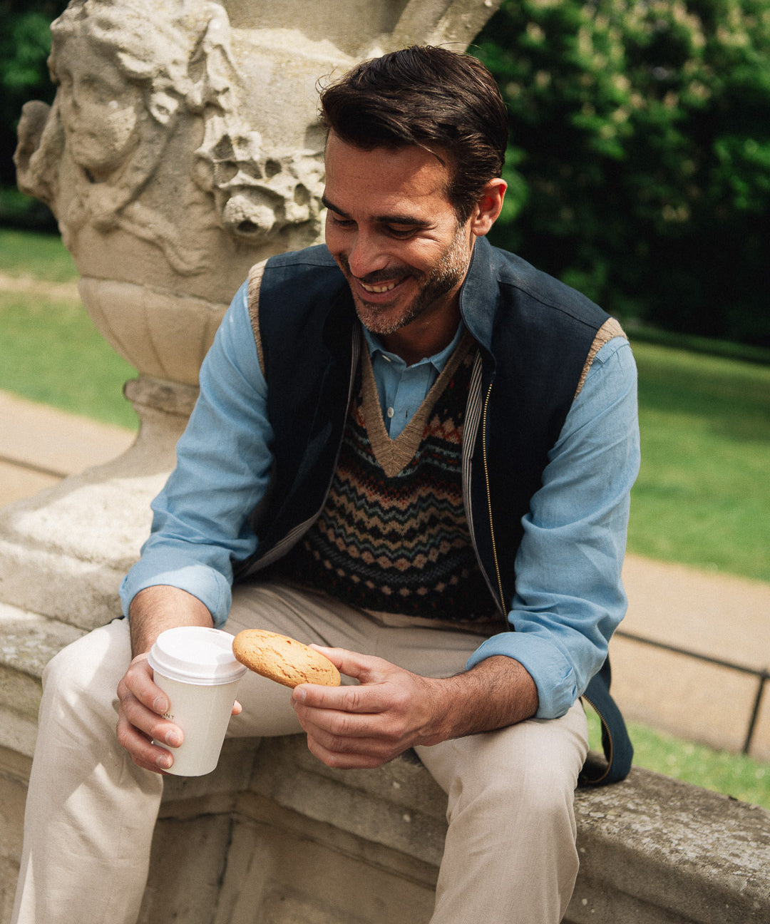 Man sitting outdoors drinking from a cup of coffee wearing a Darzi navy linen Hillgate gilet