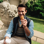 Man sitting outdoors, holding a pastry and a coffee cup, with greenery in the background, wearing a Darzi navy linen Hillgate vest