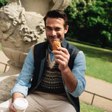Man sitting outdoors, holding a pastry and a coffee cup, with greenery in the background, wearing a Darzi navy linen Hillgate vest