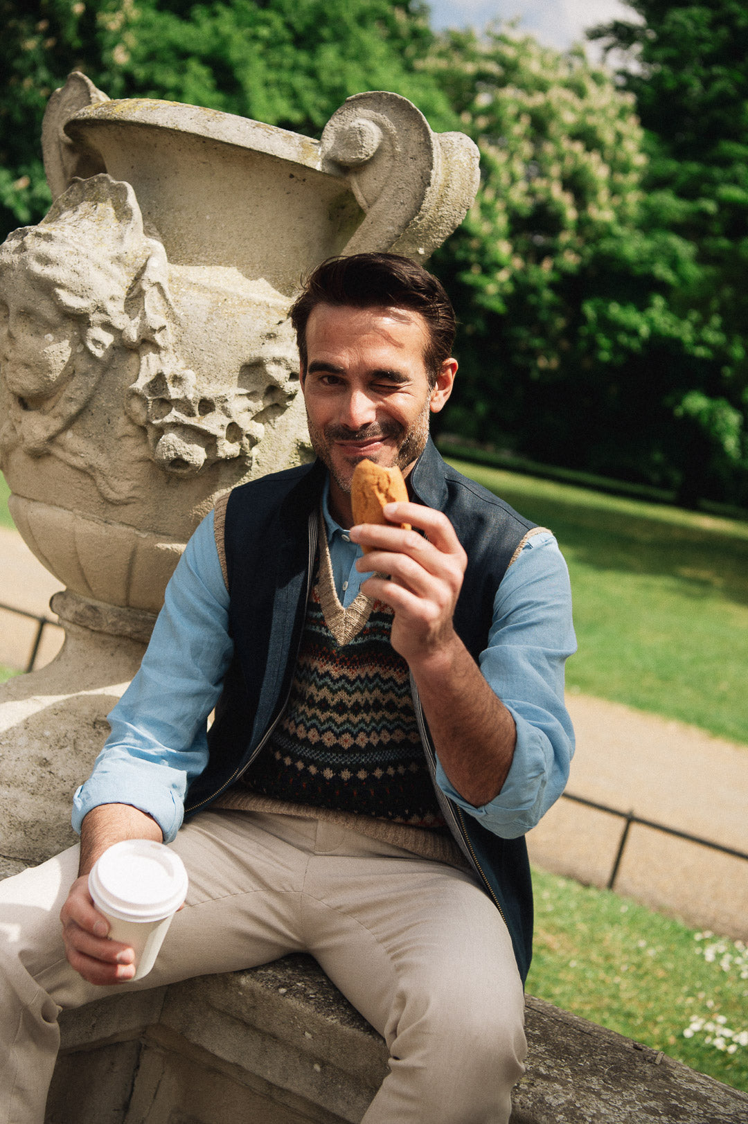 Man sitting outdoors, holding a pastry and a coffee cup, with greenery in the background, wearing a Darzi navy linen Hillgate vest