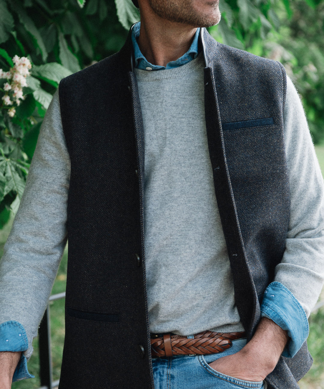 Man standing outdoors wearing a Darzi Helmsley wool waistcoat in Blue Brown Twill over a grey sweater, with jeans