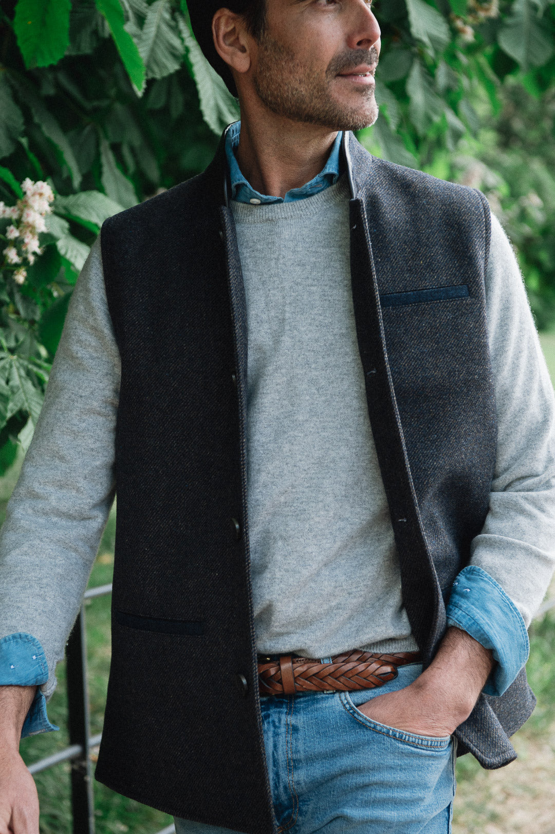 Man standing outdoors wearing a Darzi Helmsley wool waistcoat in Blue Brown Twill over a grey sweater, with jeans