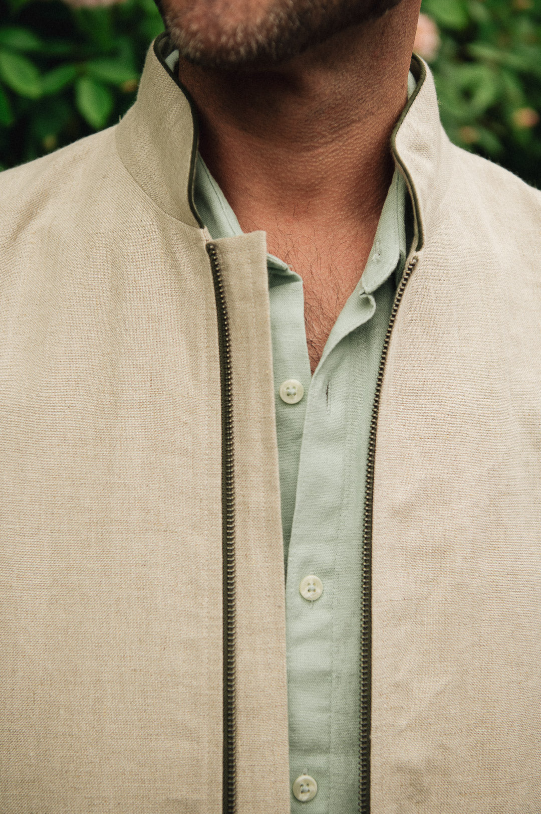 Close-up of a man wearing a Darzi limestone linen Hillgate gilet over a light green shirt with a blurred green background