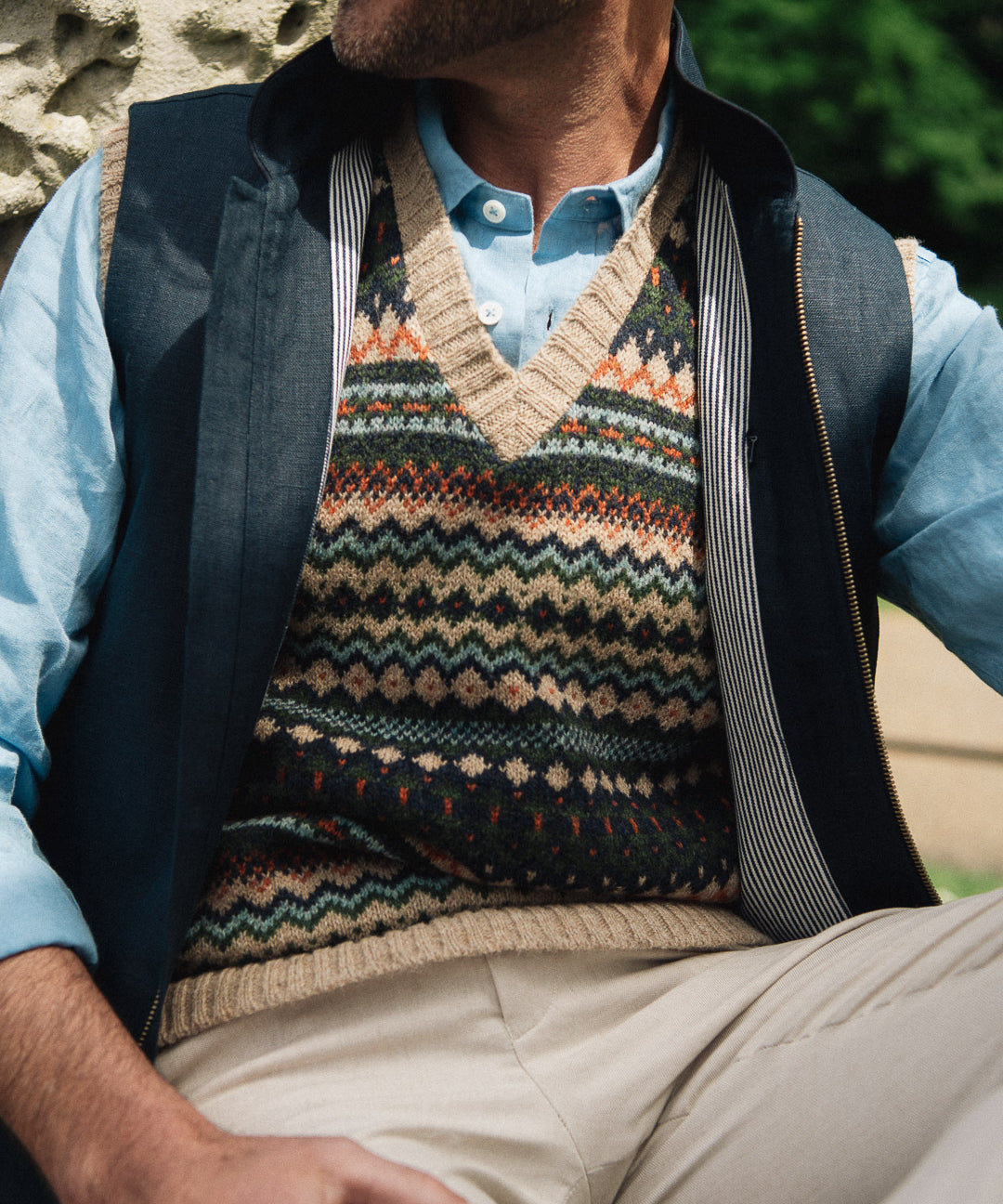 Man wearing a patterned sweater vest over a light blue shirt with beige pants, sitting outdoors wearing a Darzi navy linen Hillgate gilet