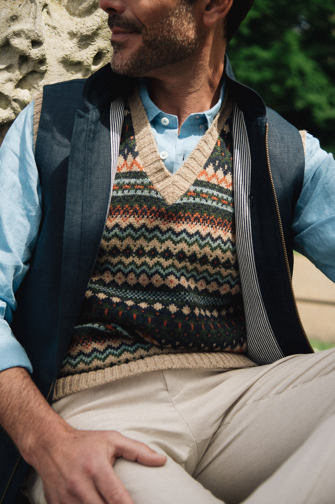 Man wearing a patterned sweater vest over a light blue shirt with beige pants, sitting outdoors wearing a Darzi navy linen Hillgate gilet