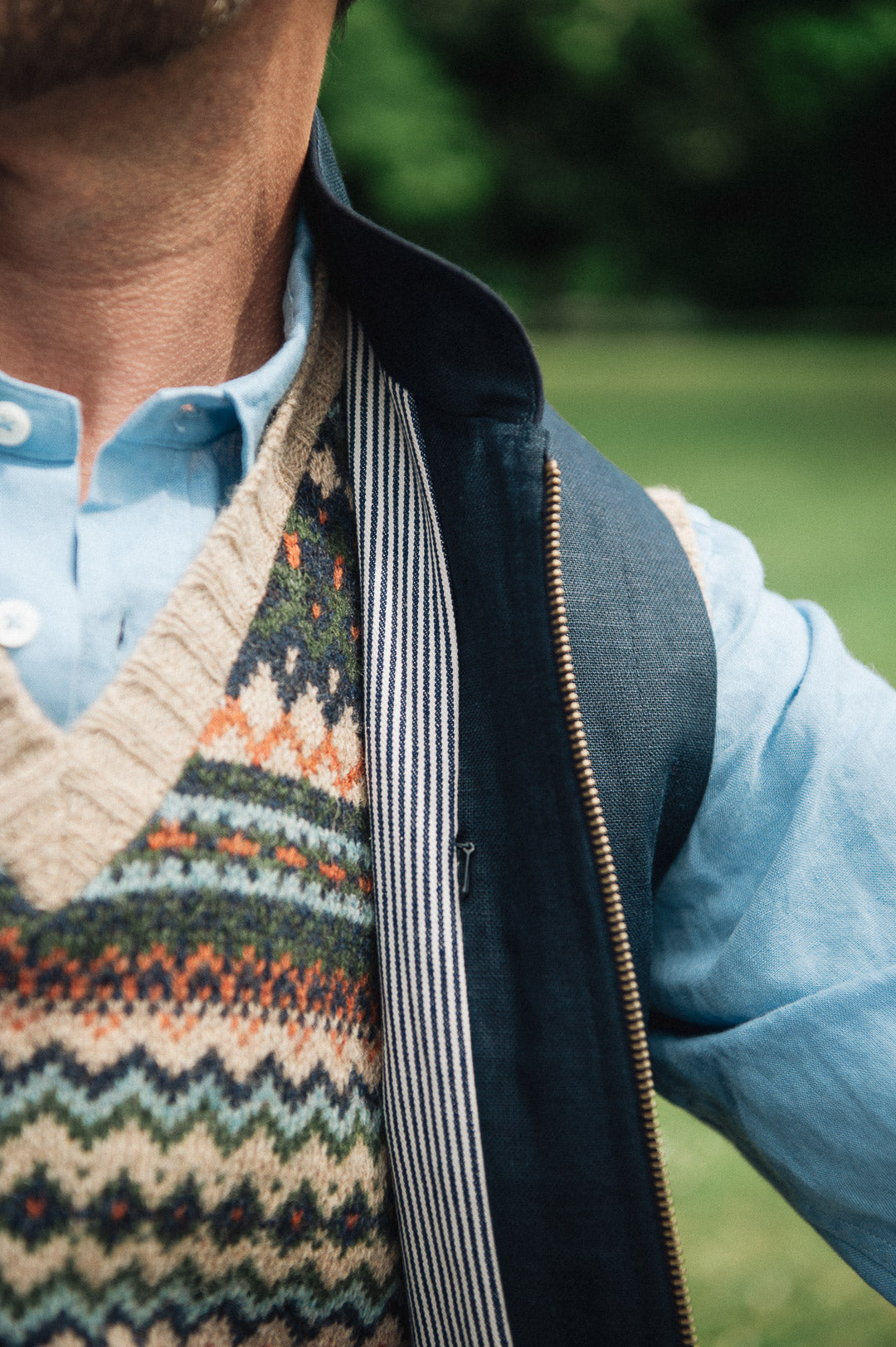 Person wearing a patterned jumper over a light blue shirt, under a Darzi navy linen Hillgate gilet with a blurred green background
