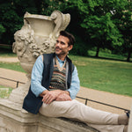 Man sitting on a stone bench in a park with trees and greenery in the background wearing Darzi navy linen Hillgate gilet