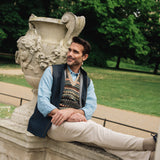 Man sitting on a stone bench in a park with trees and greenery in the background wearing Darzi navy linen Hillgate gilet