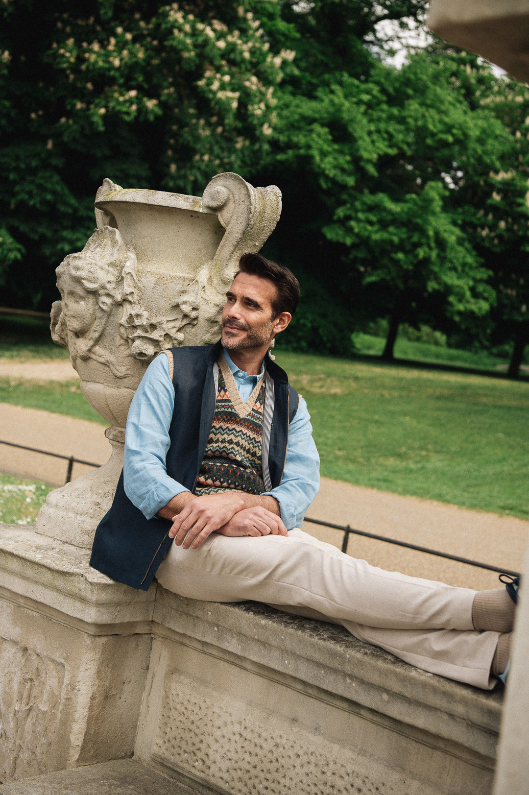 Man sitting on a stone bench in a park with trees and greenery in the background wearing Darzi navy linen Hillgate gilet
