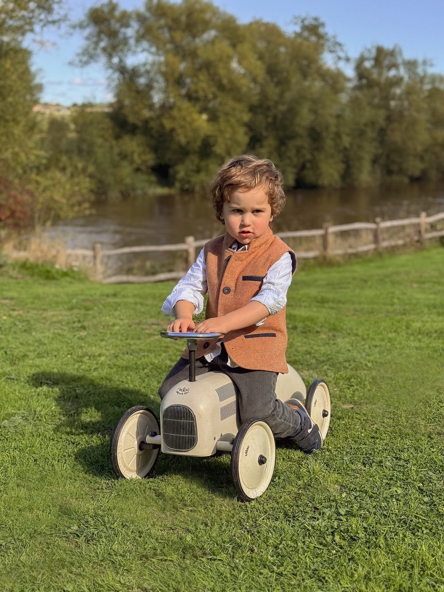 Boy wearing Little Darzi wool gilet in Coral Red sitting on toy car